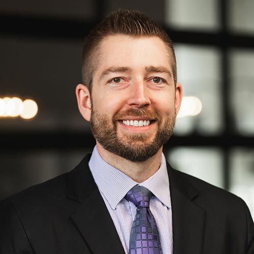 Alex R. Schoephoerster; A man with short brown hair and beard, wearing a black suit, light shirt, and purple tie, smiles against a blurred office background with soft lighting.