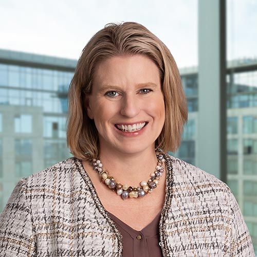 Kirstin D. Kanski; A woman with short, light brown hair smiles warmly. She wears a tweed jacket and pearl necklace, standing in front of a modern office window. The mood is professional and approachable.