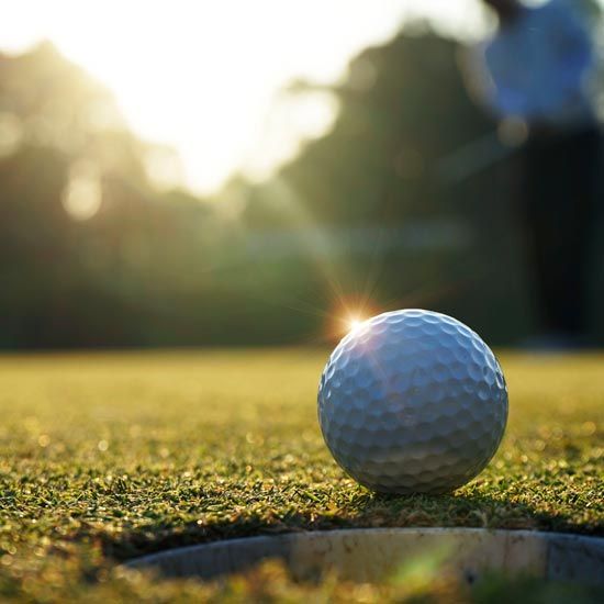 Portfolio-Management-FEATURE - Closeup of a golf ball sitting within an inch of the hole while the sun rises in the background glinting off the edge of the ball.